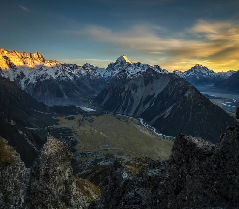 A high-altitude view of a person standing on a rocky outcrop overlooking a vast glacial valley, with the sun-drenched peak of Aoraki / Mount Cook in the distance under a golden sky. Credit: Mackenzie Tourism - Lee Cook
