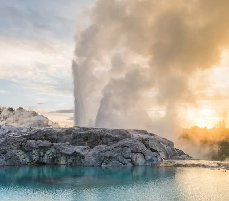 The Pōhutu Geyser erupts with a massive plume of steam and water behind a rocky silica terrace, reflected in a calm turquoise pool under a golden sky.