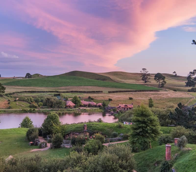Scenic Hobbiton Movie Set with green hills, lake, trees, and buildings at sunset.