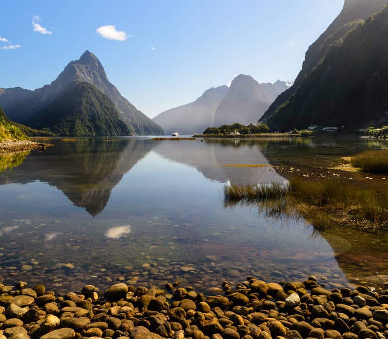 Milford Sound calm water reflecting mountainous landscape with rocky shore under blue sky.