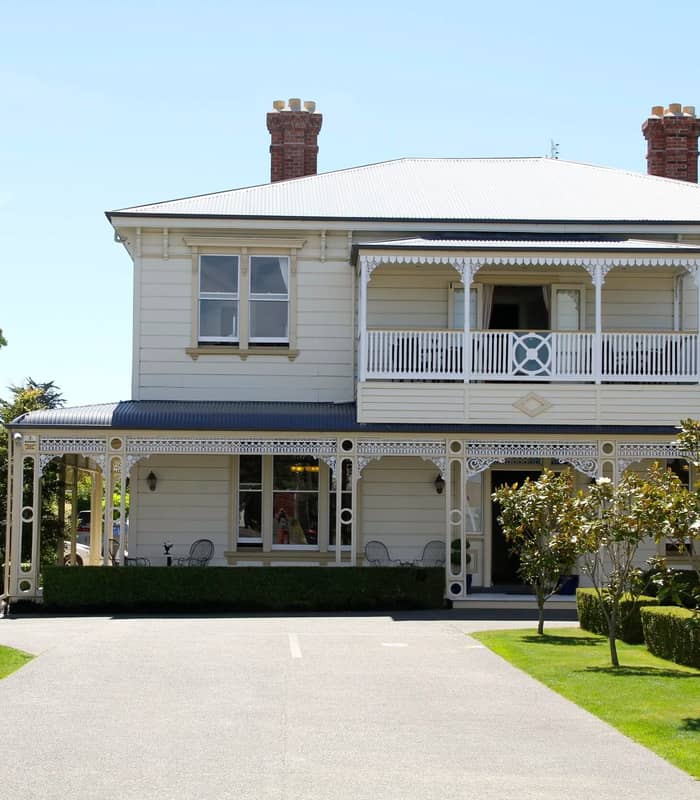 Front view of Merivale Manor House with balcony, veranda, trees, and garden on a sunny day.