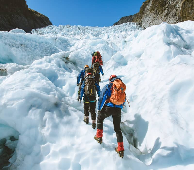 Group hiking up a glacier with crampons and backpacks on a sunny day.