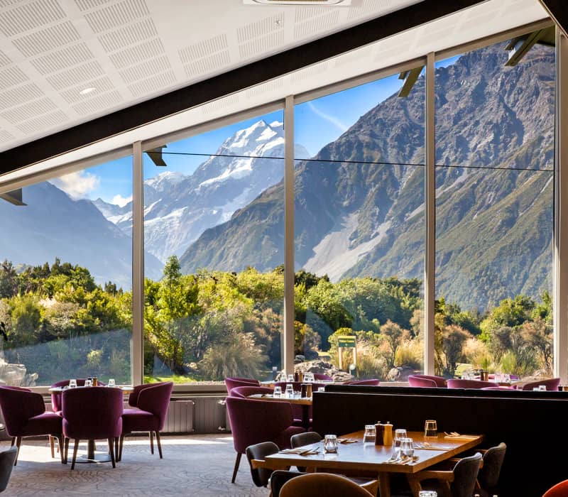 Indoor dining room with purple chairs and large windows showing alpine mountains and greenery outside.