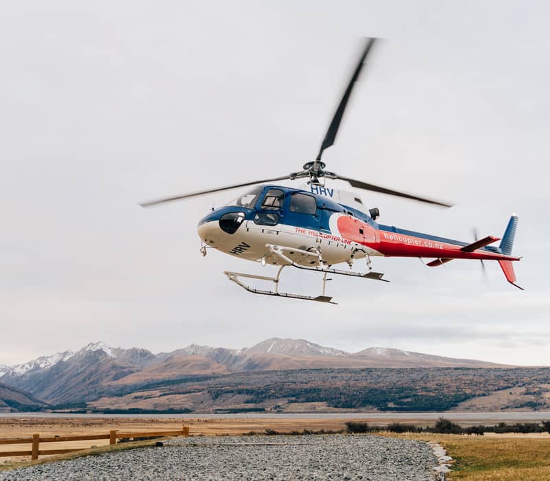 Helicopter flying above rocky and grassy land with snowy mountains in the background under cloudy sky.