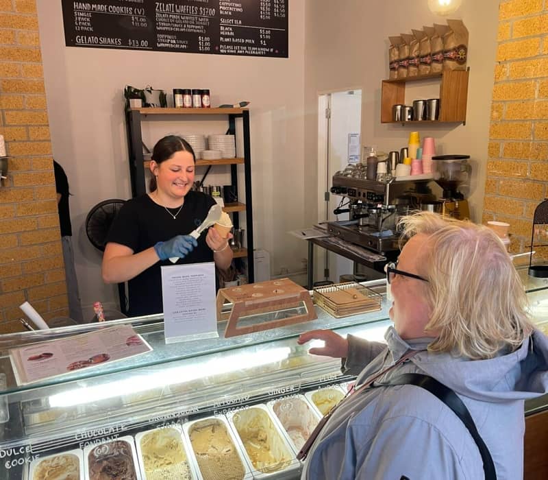 Server scooping gelato for customer at an indoor service counter with menu and coffee machine.