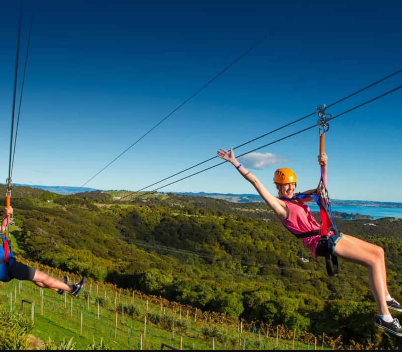 Two people ziplining over green hills with blue sky and coastline in the background.