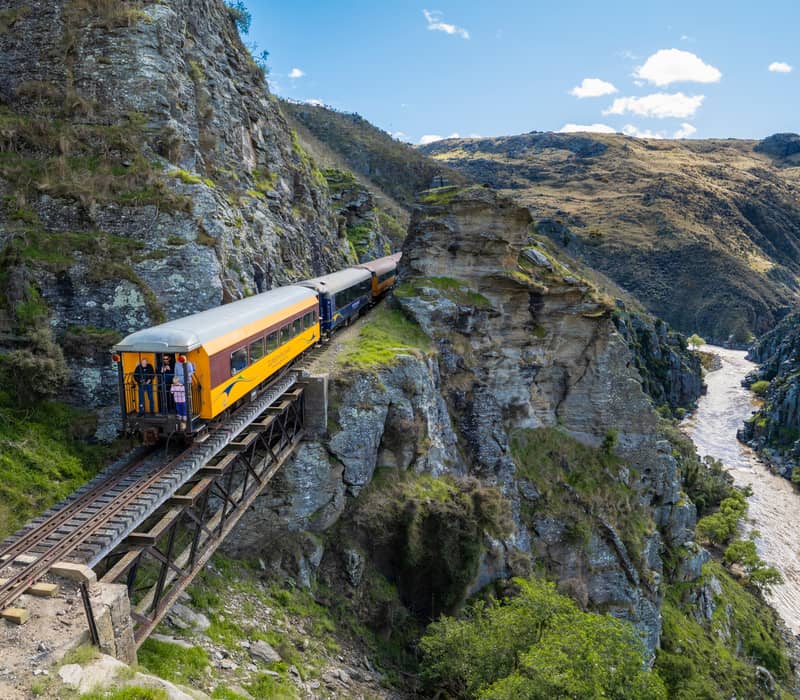 Passenger train crossing a narrow railway bridge over a gorge with river and rocky mountain landscape under blue sky.