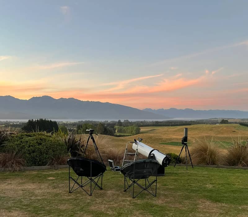 Telescopes and chairs set on grass overlooking mountains at sunset with a colorful sky.