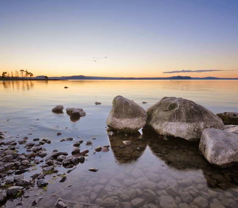 Sunset over Lake Taupo with rocks in the water and calm reflections.