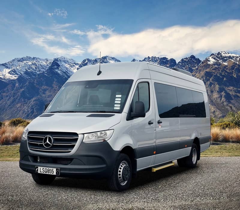 A pristine silver Mercedes-Benz Sprinter executive van parked on an asphalt turnaround, showcased against a majestic backdrop of the rugged, snow-dusted Remarkables mountain range under a clear blue sky. Credit: Limousine Services Queenstown