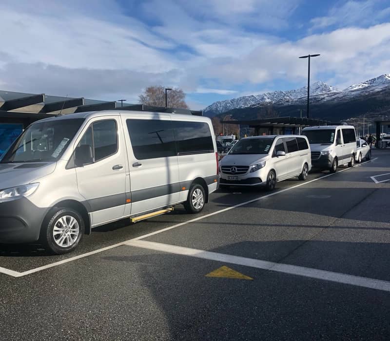 A line of premium silver Mercedes-Benz passenger vans parked in the commercial transport lane at Queenstown Airport, with the snow-capped peaks of The Remarkables mountain range towering under a cloudy blue sky.