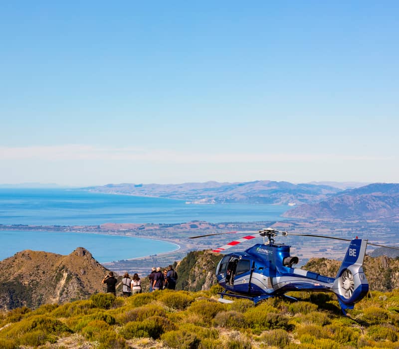 Tourists by a blue helicopter on mountain overlooking coastal landscape and ocean.
