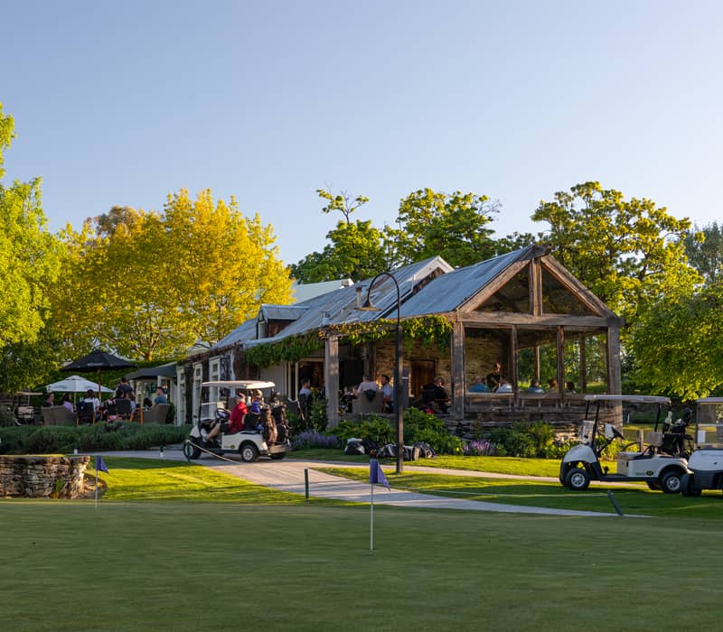 Cafe building with outdoor seating, golf carts, and green golf course under clear sky.