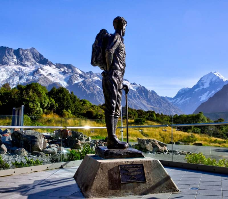 Statue of a man with backpack stands against a backdrop of snow-capped mountains and clear blue sky.