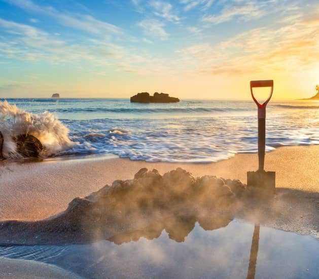 Shovel in sand on beach with sunrise, waves, and rocks nearby.