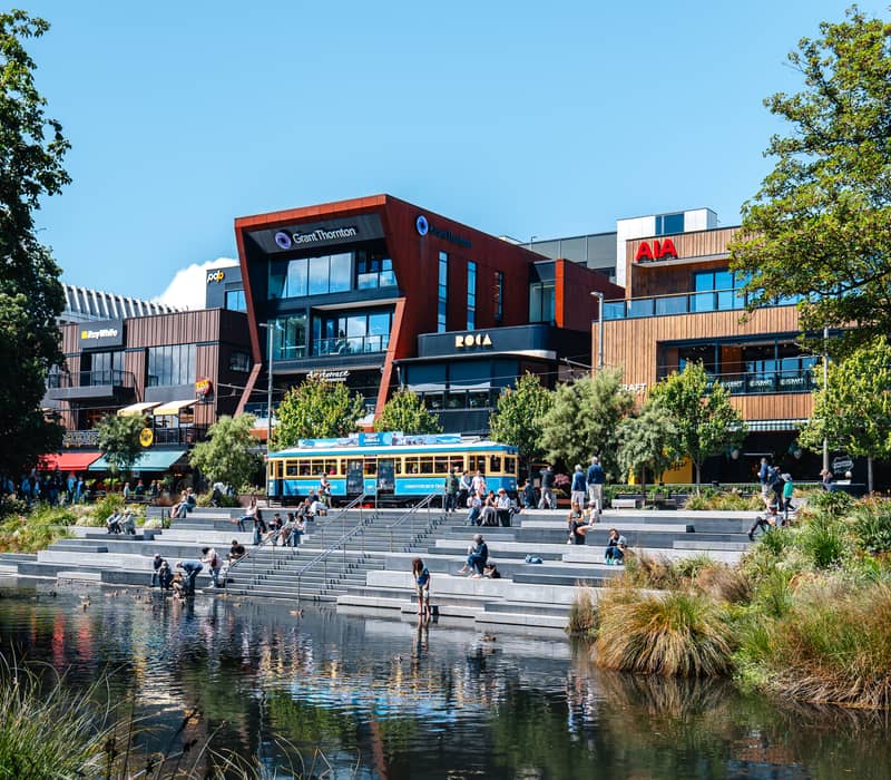 People sitting on concrete steps by a river with modern buildings and a blue tram in the background.