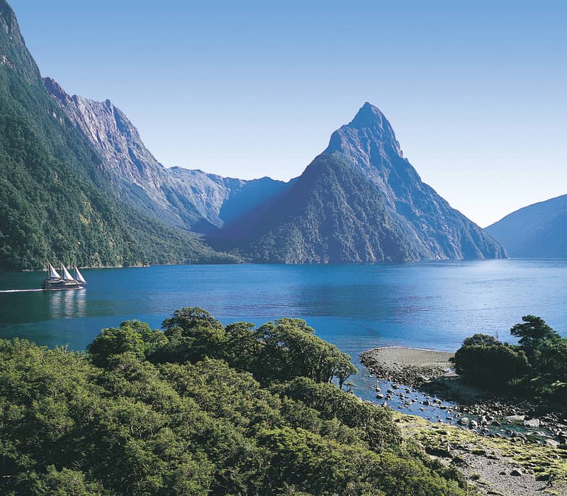 Sailboat on calm blue water surrounded by towering green mountains under clear sky.