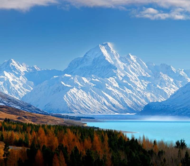 Snow-covered mountain peaks above a turquoise lake with mixed forest in foreground under a blue sky.