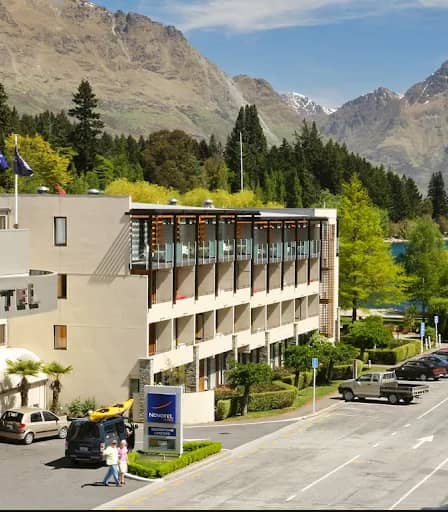 Modern hotel building with balconies, parked cars, mountain and lake scenery under clear sky.
