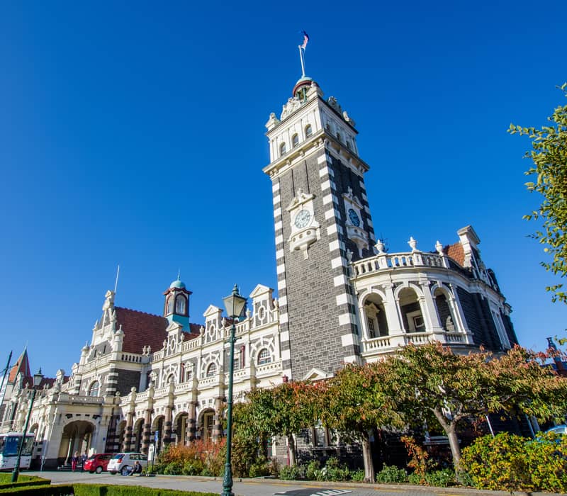 A low-angle, sunny view of the historic Flemish Renaissance-style Dunedin Railway Station in New Zealand, featuring its prominent dark basalt and white limestone clock tower under a clear blue sky. Credit: Grace Thang - Shutterstock