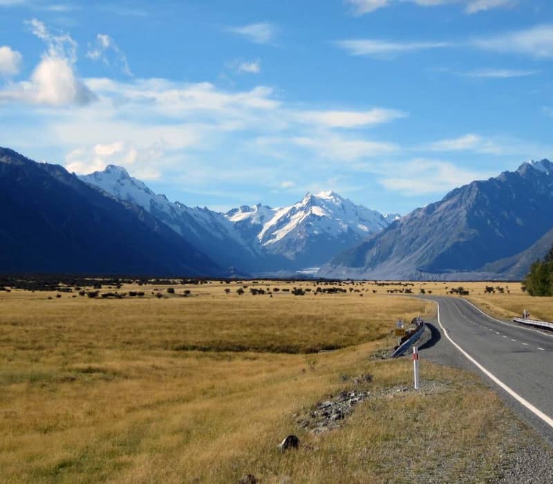 Road through grassland valley with snow-capped mountains under a blue sky with clouds.