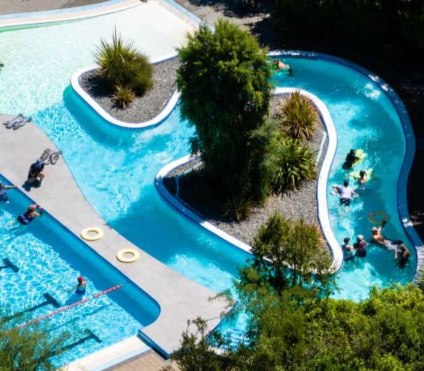 Aerial view of Hanmer Springs Thermal Pools with people swimming and relaxing in blue water pools surrounded by trees.