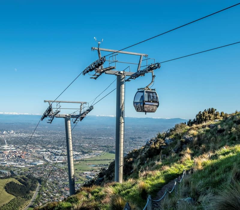 Gondola cable car over mountain with city and snow-capped peaks in the background under blue sky