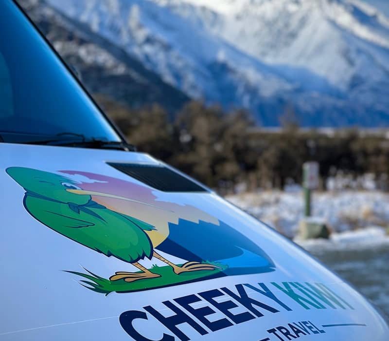 Cheeky Kiwi Travel van with a kiwi logo in front of snowy Mt Cook mountains in winter.