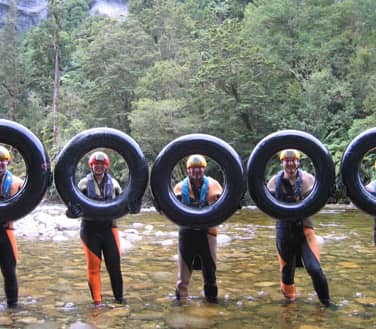 Five people in wetsuits holding inner tubes standing in a shallow river with forest in the background.