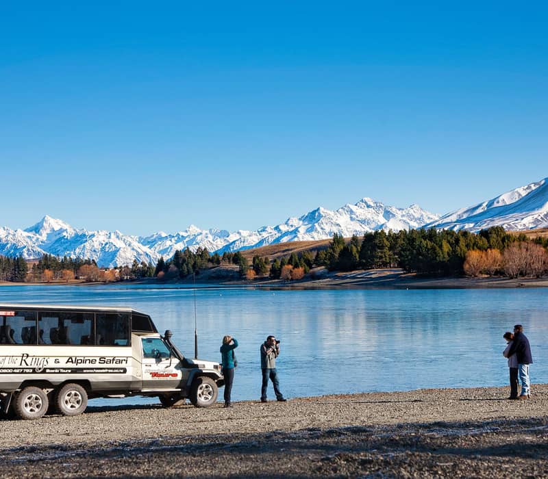 Tour vehicle by lake with snowy mountains and people taking photos and enjoying the view.