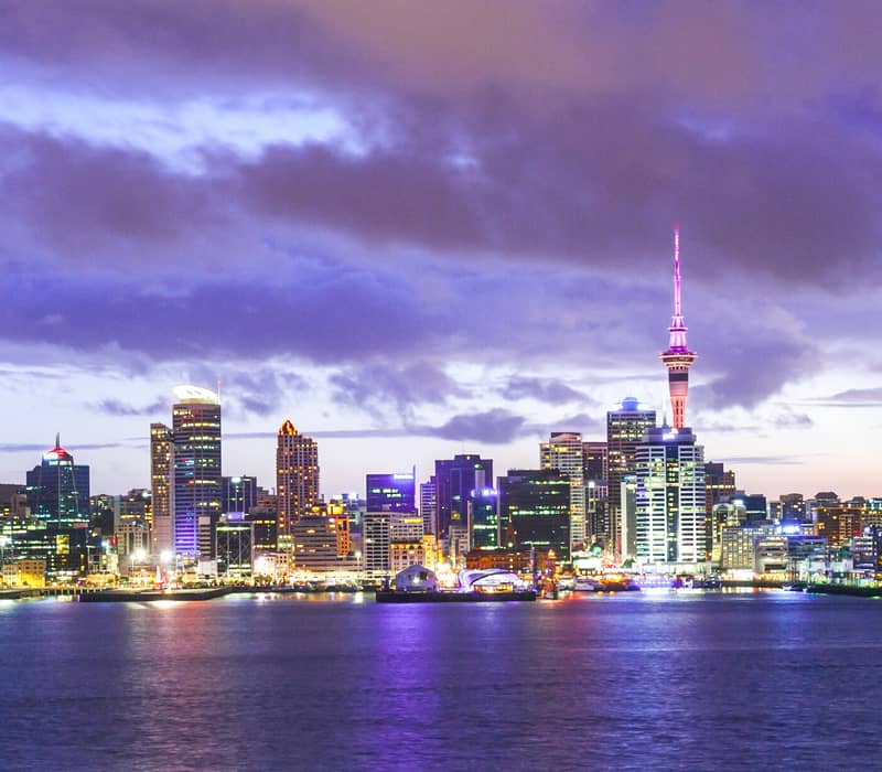 Auckland city skyline at twilight with purple clouds and city lights reflecting on the harbour water.