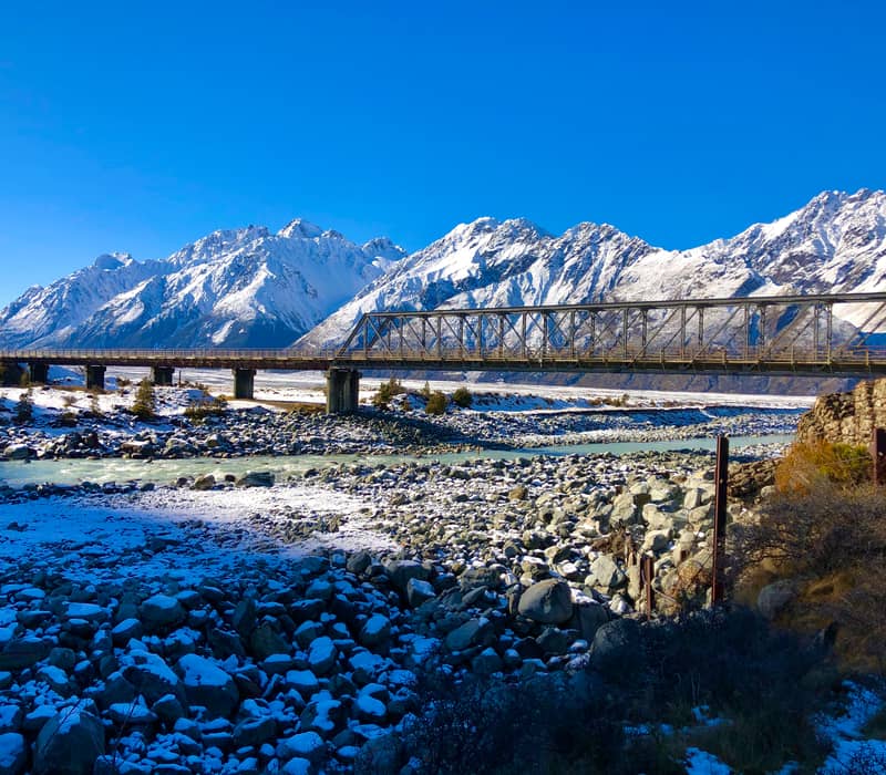 Metal bridge over a river with snow-covered rocks and mountains under a blue sky.