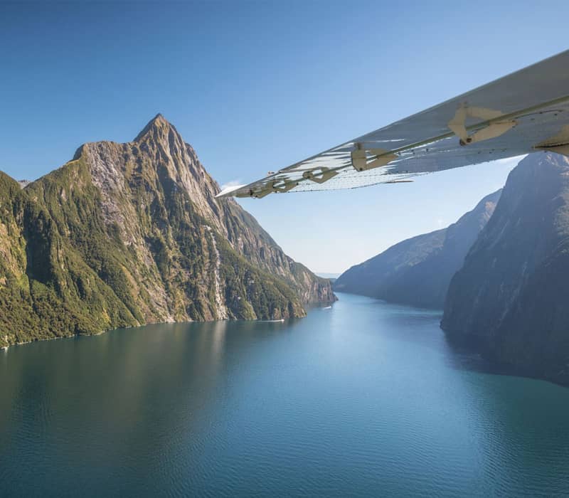 Aerial view of Milford Sound with mountains, water, cruise boats, and airplane wing under clear sky.