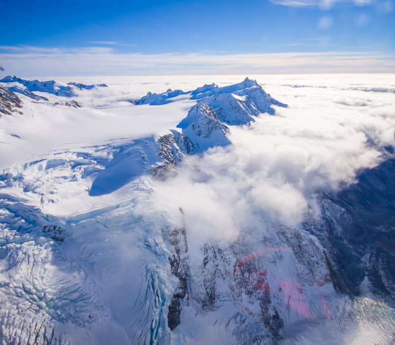 An aerial view of the Southern Alps' snow-capped peaks and glaciers partially covered by a thick blanket of white clouds under a clear blue sky. Credit: Fine Tours Group