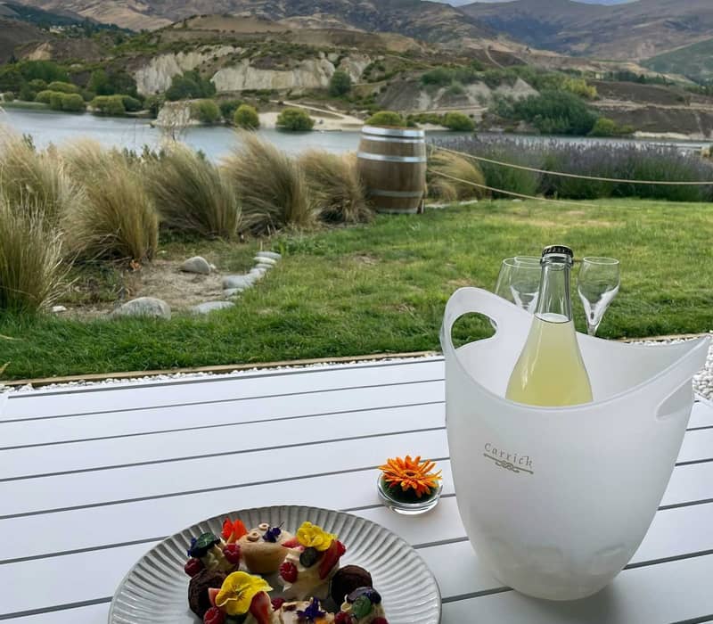 Outdoor table at Carrick Winery with petit fours, a cocktail in an ice bucket, and mountains and lake in the background.
