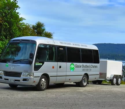 White Glacier Shuttles & Charters bus with trailer parked by a lake with trees and hills in background.