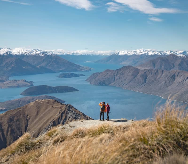 Two hikers on Roy's Peak with a wide view of Lake Wanaka and snowy mountains in the background.