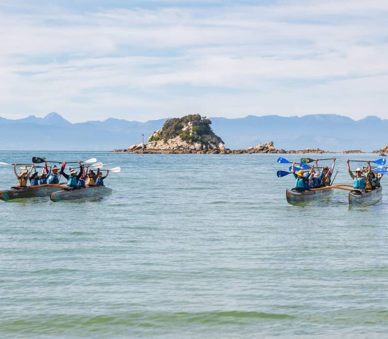 Two groups kayak in double canoes on calm sea near small islands, mountains in background.