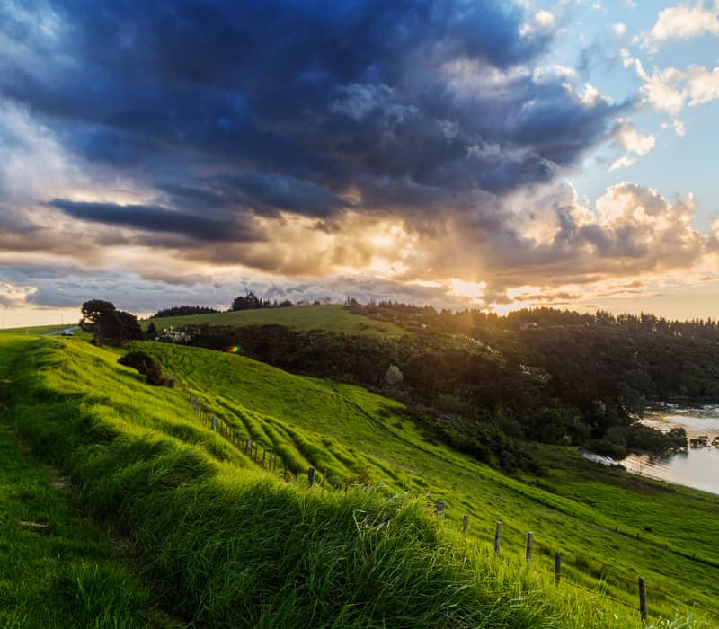Lush green hills with a wooden fence overlooking a calm bay under a sky with dark clouds and sunbeams.