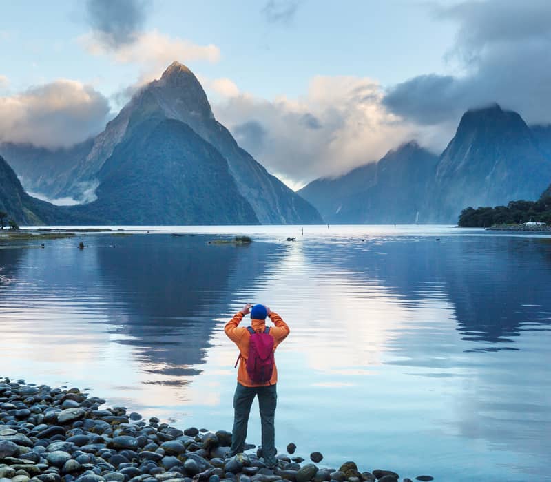 A person in an orange coat looking out over Milford Sound with water and hills in the background