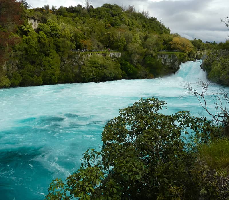 Powerful, turquoise-blue water surges over Huka Falls into a wide, churning pool surrounded by lush green native forest under a cloudy sky.