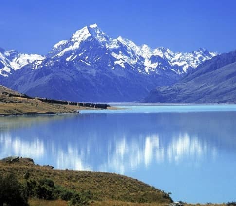 Snow-covered Mount Cook reflected in a peaceful lake with grassy hills in foreground under clear blue sky.