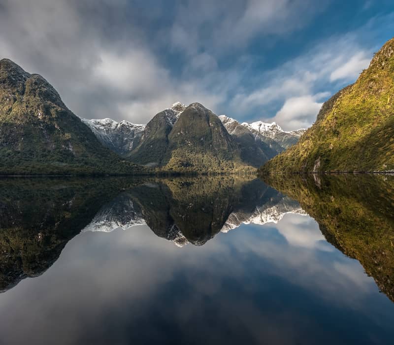 Mountain peaks reflecting on calm lake water with partly cloudy sky above.