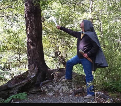 Person in medieval cloak holding sword standing on tree roots in forest.