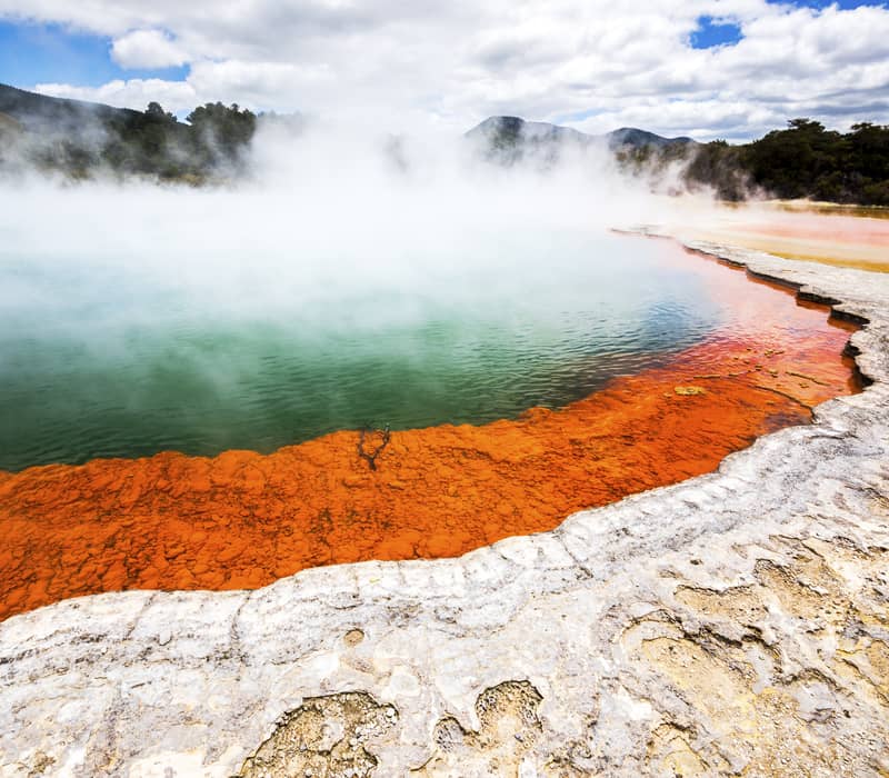 Steaming turquoise water fills the Champagne Pool geothermal spring, bordered by a vibrant orange mineral edge and white silica crust at Wai-O-Tapu.