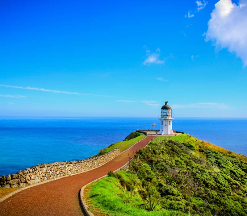 White lighthouse atop a green cliff with a pathway leading to it under a bright blue sky.
