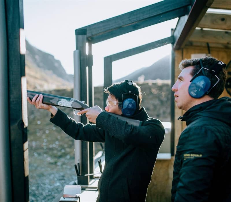 Two men at outdoor shooting range with ear protection and shotgun, mountains in background.