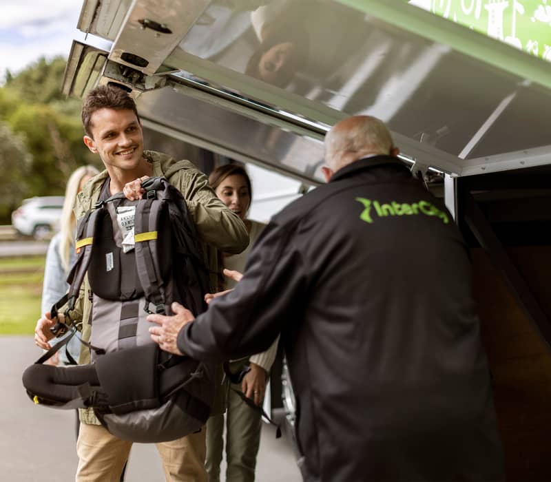 Young man handing backpack to bus worker for loading into luggage compartment outdoors.