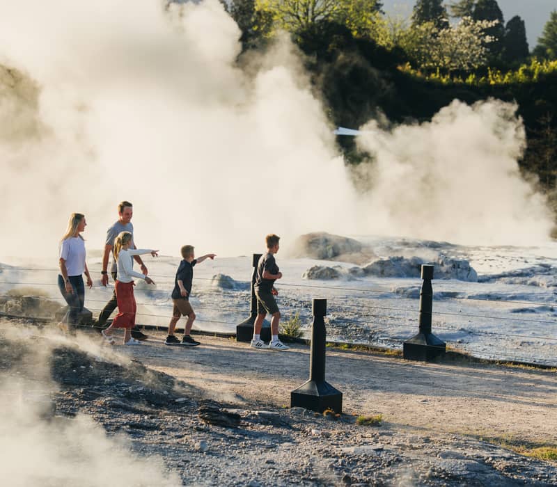 Family walking along a pathway with geothermal steam rising nearby in a green outdoor area.