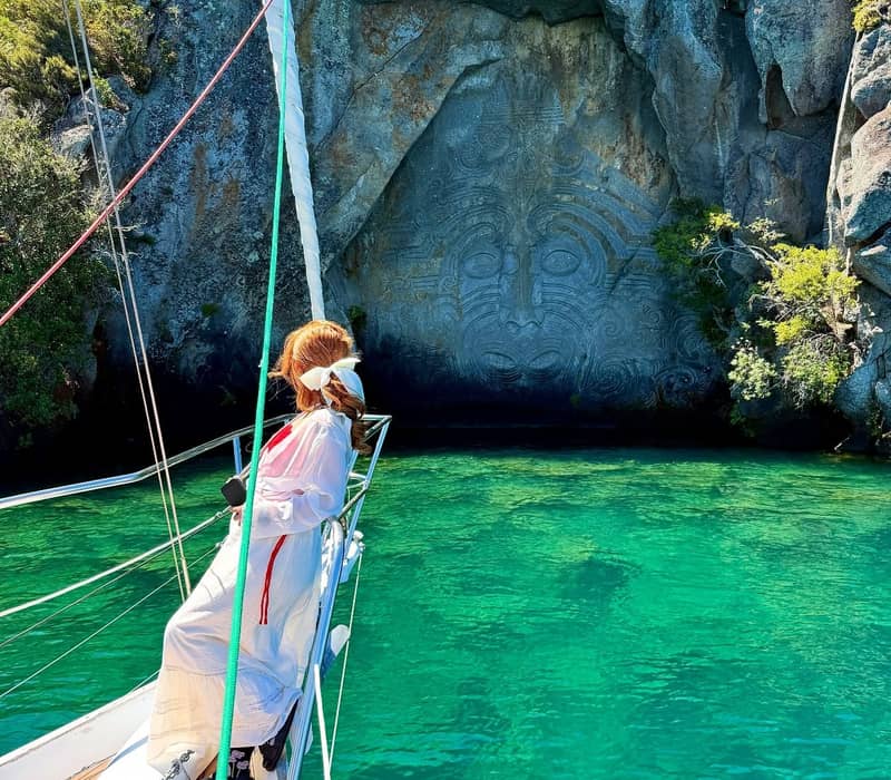 Woman on sailboat looking at rock carving in cave with clear green water and trees.